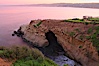 A large natural sea cave lies below a sandstone bluff in La Jolla at sunrise with a pink sky, Black's Beach in the distant. La Jolla, California, USA. Image #20250