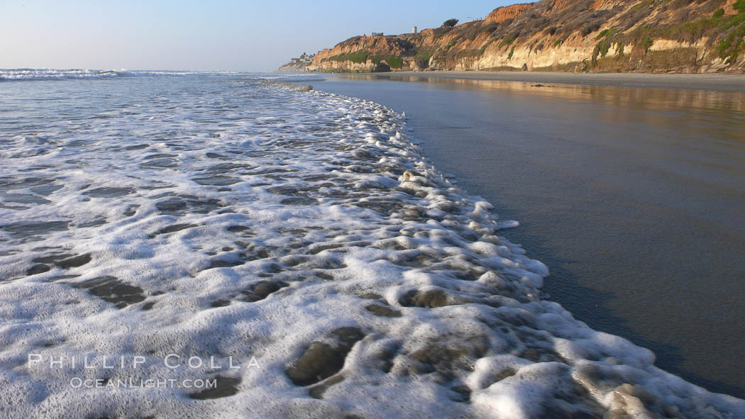 Ocean water washes over a flat sand beach, sandstone bluffs rise in the background, sunset, Carlsbad, California