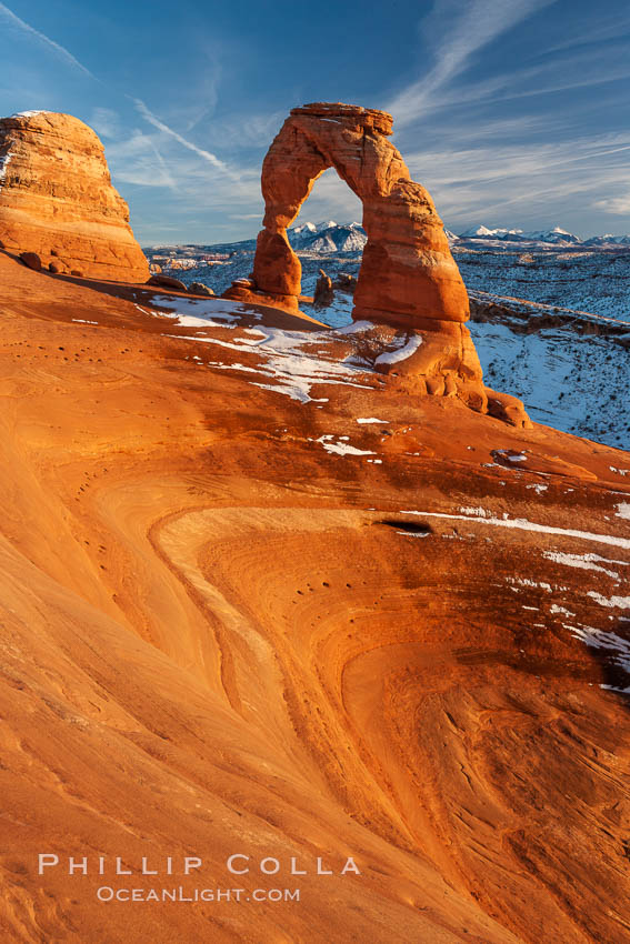 Delicate Arch, dusted with snow, at sunset, with the snow-covered La Sal mountains in the distance.  Delicate Arch stands 45 feet high, with a span of 33 feet, atop of bowl of slickrock sandstone. Arches National Park, Utah, USA, natural history stock photograph, photo id 18110