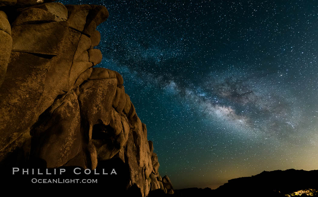 The Milky Way rises above a huge wall of stone, stars fill the night sky and soar over the distant lights of campers, Joshua Tree National Park, California