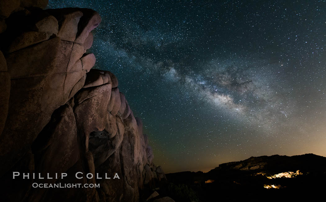 The Milky Way rises above a huge wall of stone, stars fill the night sky and soar over the distant lights of campers. Joshua Tree National Park, California, USA, natural history stock photograph, photo id 27899