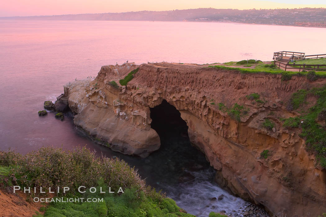 A large natural sea cave lies below a sandstone bluff in La Jolla at sunrise with a pink sky, Black's Beach in the distant