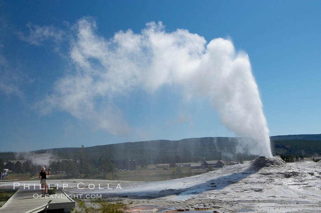 A visitor videotapes the eruption of Lion Geyser, with Old Faithful Inn visible in the distance. Lion Geyser, whose eruption is preceded by a release of steam that sounds like a lion roaring, erupts just once or a few times each day, reaching heights of up to 90 feet. Upper Geyser Basin, Yellowstone National Park, Wyoming