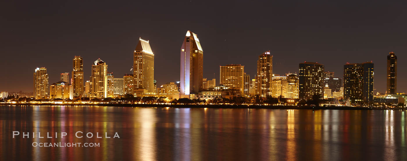 San Diego city skyline at night, showing the buildings of downtown San Diego reflected in the still waters of San Diego Harbor, viewed from Coronado Island. A panoramic photograph, composite of seven separate images