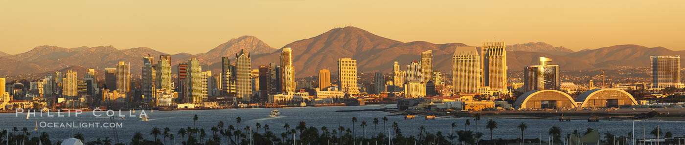 San Diego city skyline, showing the buildings of downtown San Diego rising above San Diego Harbor, viewed from Point Loma at sunset, with mountains of the Cleveland National Forest rising in the distance. A panoramic photograph, composite of six separate images