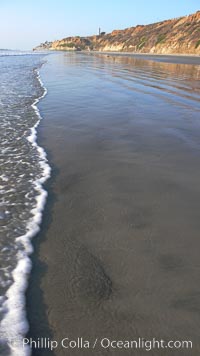 Ocean water washes over a flat sand beach, sandstone bluffs rise in the background, sunset, Carlsbad, California