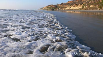 Ocean water washes over a flat sand beach, sandstone bluffs rise in the background, sunset, Carlsbad, California