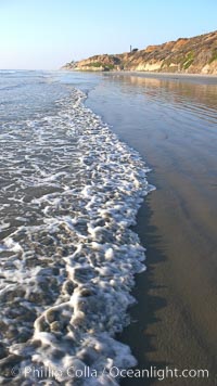 Ocean water washes over a flat sand beach, sandstone bluffs rise in the background, sunset, Carlsbad, California