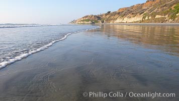 Ocean water washes over a flat sand beach, sandstone bluffs rise in the background, sunset, Carlsbad, California