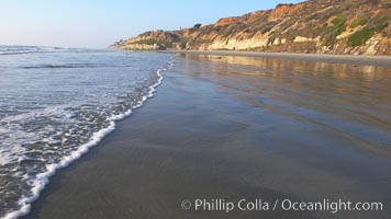 Ocean water washes over a flat sand beach, sandstone bluffs rise in the background, sunset, Carlsbad, California