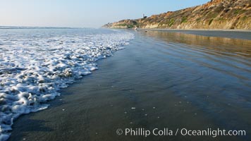 Ocean water washes over a flat sand beach, sandstone bluffs rise in the background, sunset, Carlsbad, California