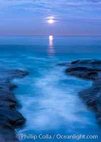 Breaking waves crash upon a rocky reef under the light of a full moon.