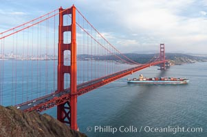 A container ship leaves San Francisco Bay, passing under the Golden Gate Bridge, viewed from the Marin Headlands with the city of San Francisco in the distance.  Late afternoon.