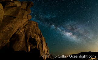 The Milky Way rises above a huge wall of stone, stars fill the night sky and soar over the distant lights of campers.