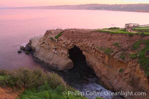 A large natural sea cave lies below a sandstone bluff in La Jolla at sunrise with a pink sky, Black's Beach in the distant.