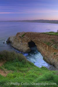 A large natural sea cave lies below a sandstone bluff in La Jolla at sunrise with a pink sky, Black's Beach in the distant