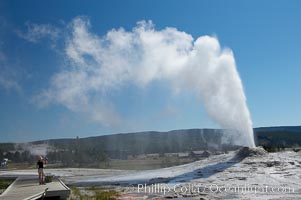 A visitor videotapes the eruption of Lion Geyser, with Old Faithful Inn visible in the distance.  Lion Geyser, whose eruption is preceded by a release of steam that sounds like a lion roaring, erupts just once or a few times each day, reaching heights of up to 90 feet.  Upper Geyser Basin, Yellowstone National Park, Wyoming