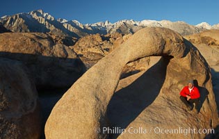 A hiker admires Mobius Arch in early morning golden sunlight, with the snow-covered Sierra Nevada Range and the Alabama Hills seen in the background, Alabama Hills Recreational Area