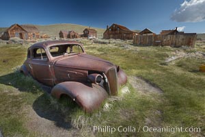 Old car lying in dirt field, Fuller Street and Green Street buildings in background.