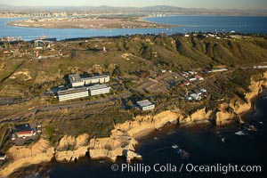 Point Loma peninsula, with scalloped sandstone cliffs edging the Pacific Ocean.  San Diego Bay, Coronado Island and the high rises of downtown San Diego are seen beyond.  Navy facilities, including SPAWAR (Space and Naval Warfare) building 600 in left center, are scattered along this section of Point Loma