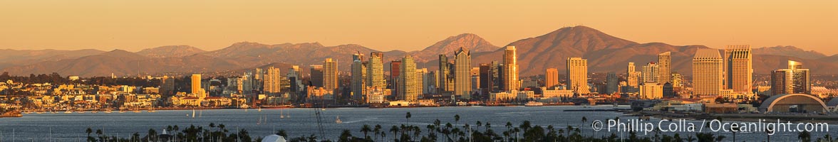 San Diego city skyline, showing the buildings of downtown San Diego rising above San Diego Harbor, viewed from Point Loma at sunset, with mountains of the Cleveland National Forest rising in the distance. A panoramic photograph, composite of seven separate images