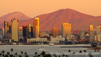 San Diego city skyline, showing the buildings of downtown San Diego rising above San Diego Harbor, viewed from Point Loma at sunset, with mountains of the Cleveland National Forest rising in the distance. Mount San Miguel is on the right and Lyons Peak to the left