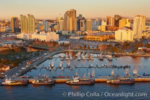 Aerial photo of San Diego Marina District, sunset, with fishing vessels docked alongside pier, Seaport Village (right) and downtown highrise office buildings rising over San Diego Bay