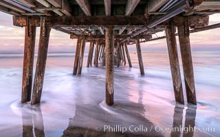 Waves break on the Imperial Beach Pier pilings, at dawn with colorful sunrise clouds over the ocean