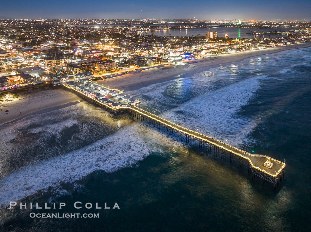 Aerial photo of Crystal Pier with Holiday Christmas Lights at night. The Crystal Pier, Holiday Lights and Pacific Ocean at sunset, waves blur as they crash upon the sand. Crystal Pier, 872 feet long and built in 1925, extends out into the Pacific Ocean from the town of Pacific Beach