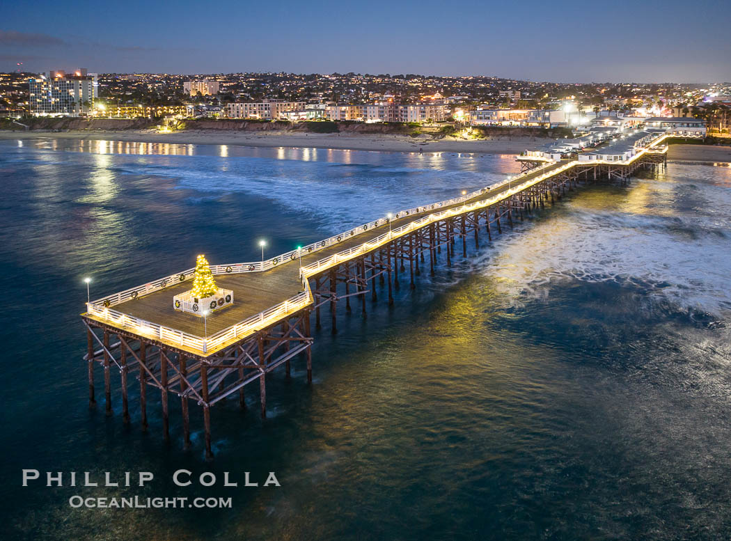 Aerial photo of Crystal Pier with Holiday Christmas Lights at night. The Crystal Pier, Holiday Lights and Pacific Ocean at sunset, waves blur as they crash upon the sand. Crystal Pier, 872 feet long and built in 1925, extends out into the Pacific Ocean from the town of Pacific Beach