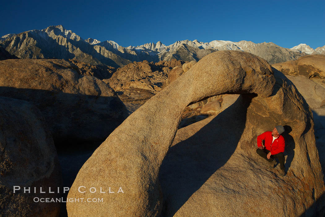 A hiker admires Mobius Arch in early morning golden sunlight, with the snow-covered Sierra Nevada Range and the Alabama Hills seen in the background. Alabama Hills Recreational Area, California, USA, natural history stock photograph, photo id 21740